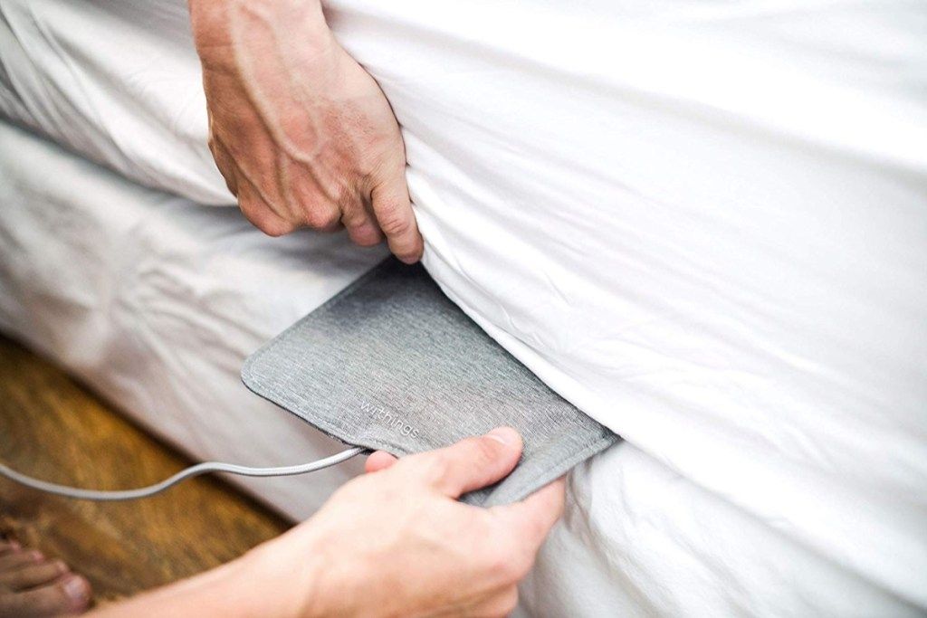 Hombre metiendo el rastreador de sueño gris debajo del colchón, lo esencial para dormir mejor