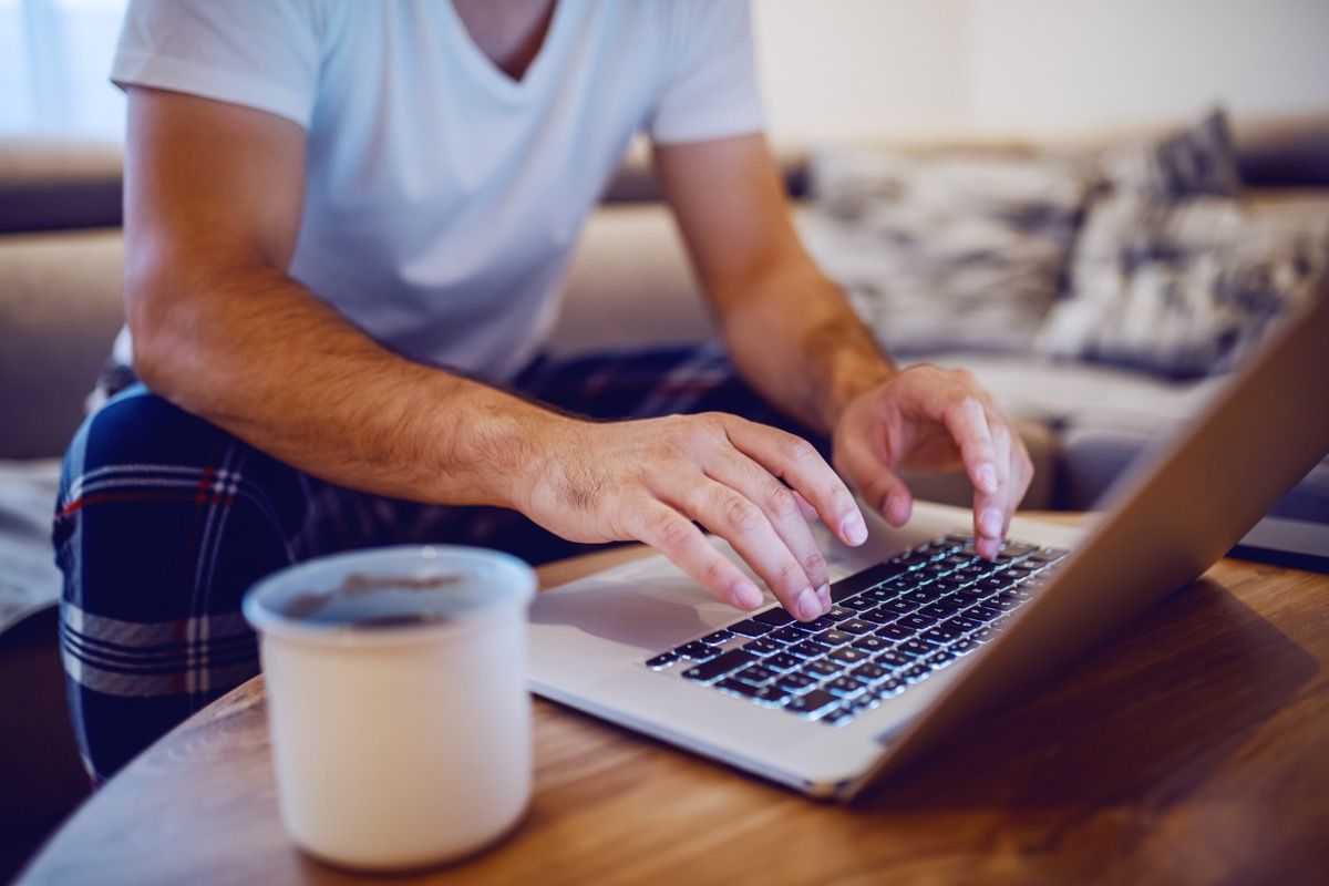 Foto lelaki yang memakai piyama di papan kekunci komputer riba dan melayari internet sambil duduk di sofa di ruang tamu pagi. Fokus selektif pada tangan.