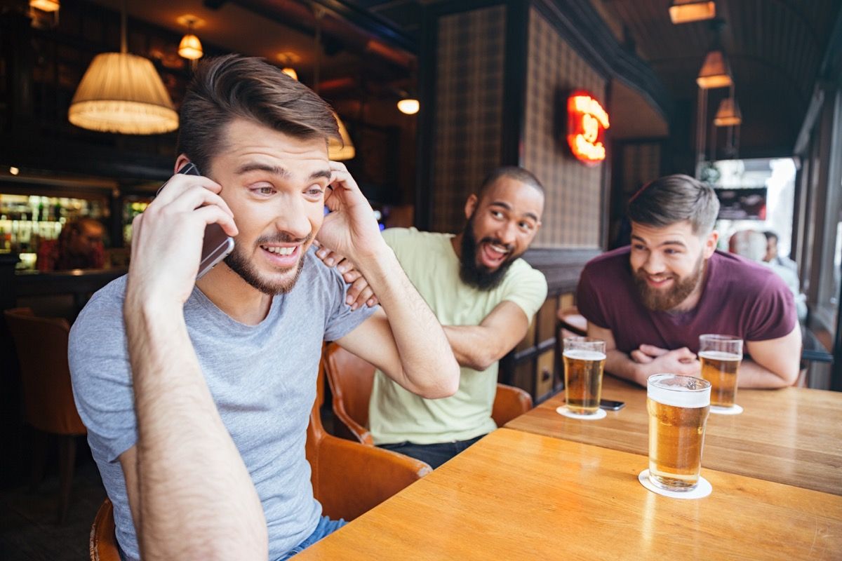 Cilvēks pa tālruni bārā ar draugiem, kas dzer alu, lietas, ko neteikt klientu apkalpošanai