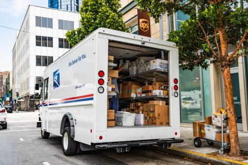   USPS の配達用バンが UPS の店舗前に停車し、Amazon の荷物を降ろしていた