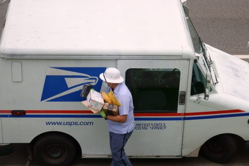   ASV pasta dienesta USPS pastnieks nēsā masku un cimdus, nesot paku kravu no pasta kravas automašīnas COVID-19 koronavīrusa pandēmijas laikā.