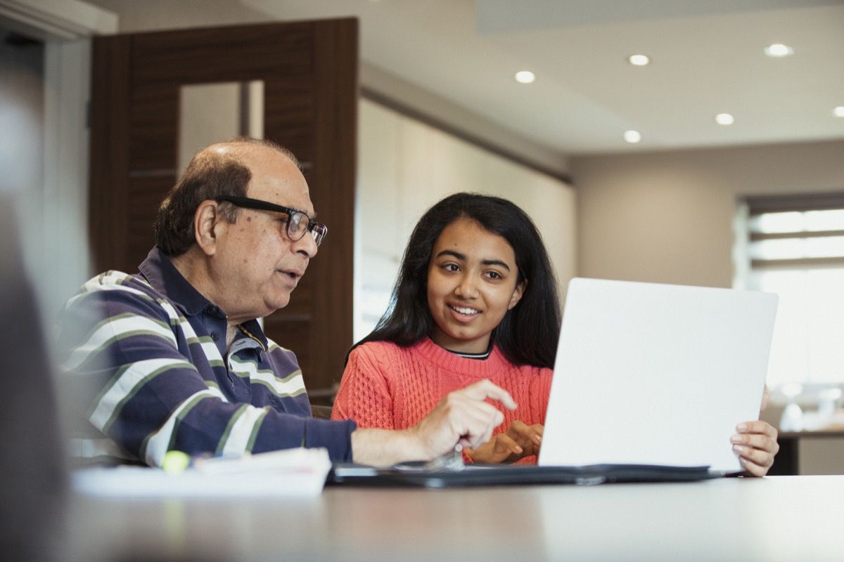 En bestefar hjelper barnebarnet sitt mens hun jobber fra en bærbar datamaskin, de ser glade ut for å være sammen og jobbe sammen.