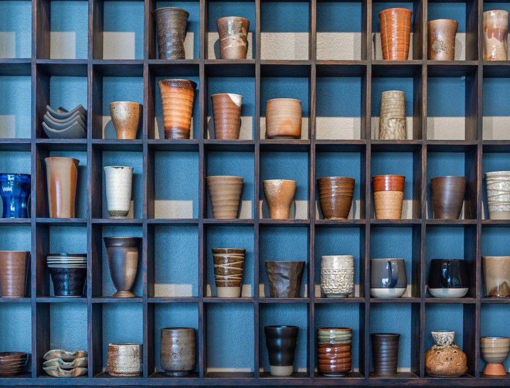 Įvairūs ir spalvingi keraminiai puodeliai ant modernios medinės lentynos mėlyname fone.