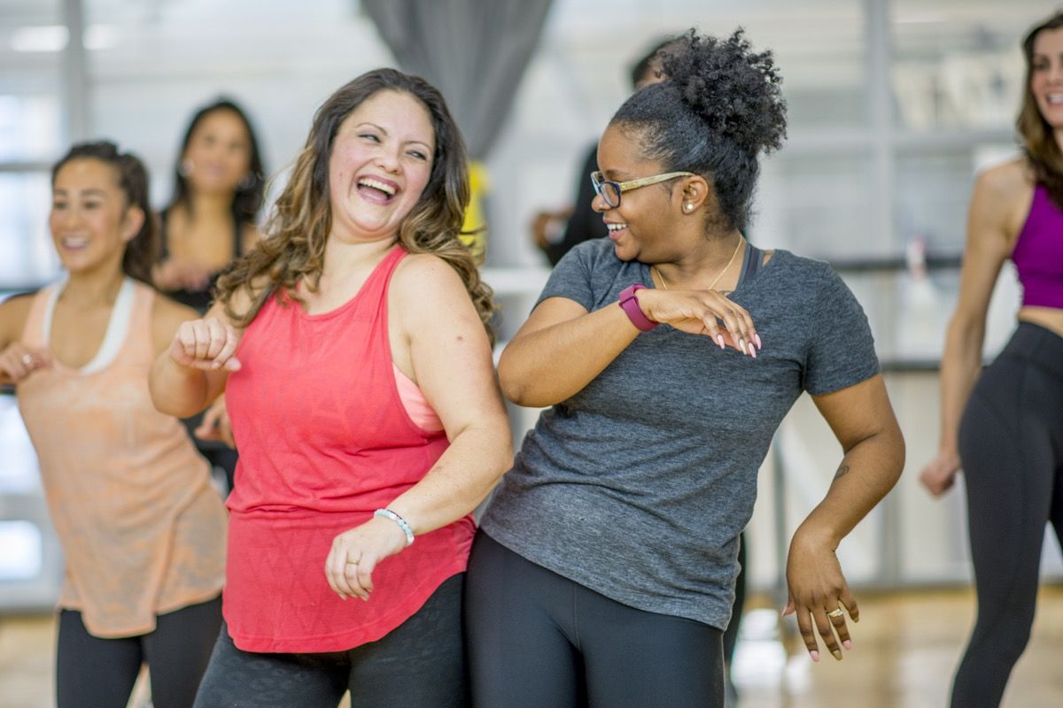 Мултиетничка група одраслих жена плеше у фитнес студију. Носе атлетску одећу. Две жене се смеју док плешу заједно.