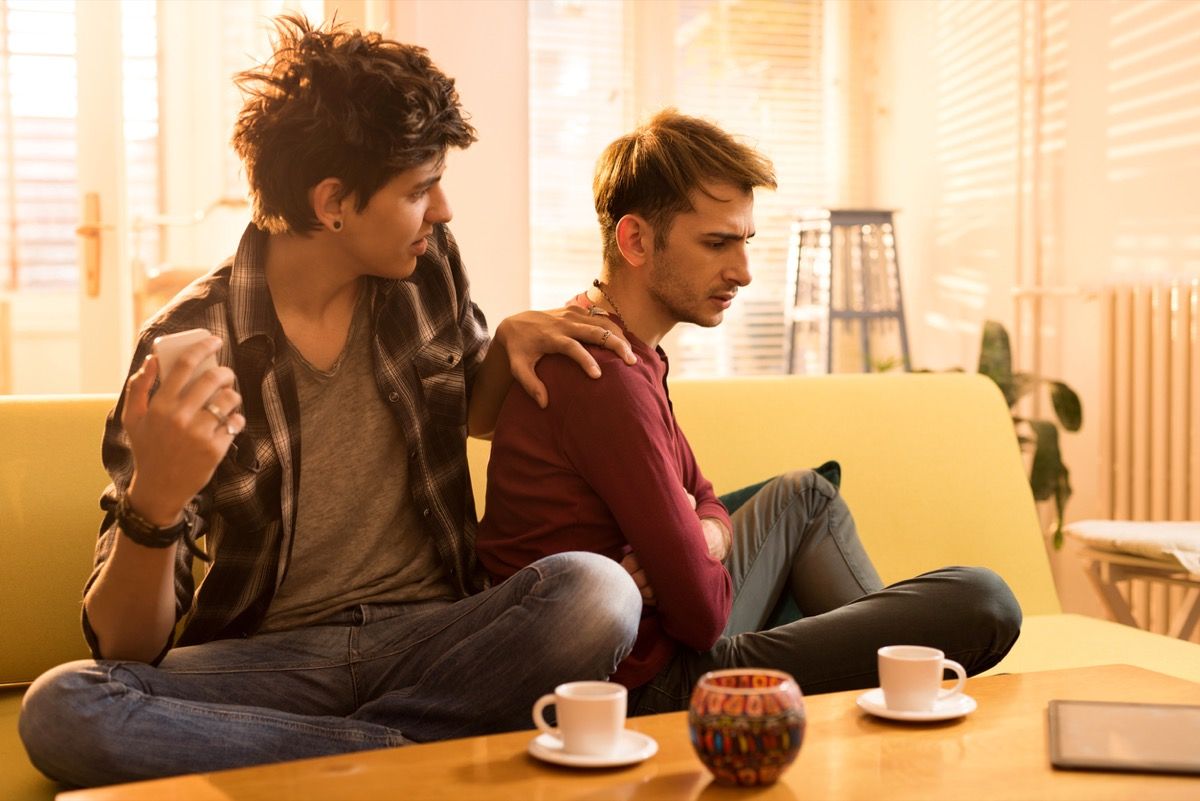 Mladý homosexuál položí ruku na rameno svojho priateľa alebo manžela, ktorý má otočený chrbtom, zatiaľ čo sedia na gauči