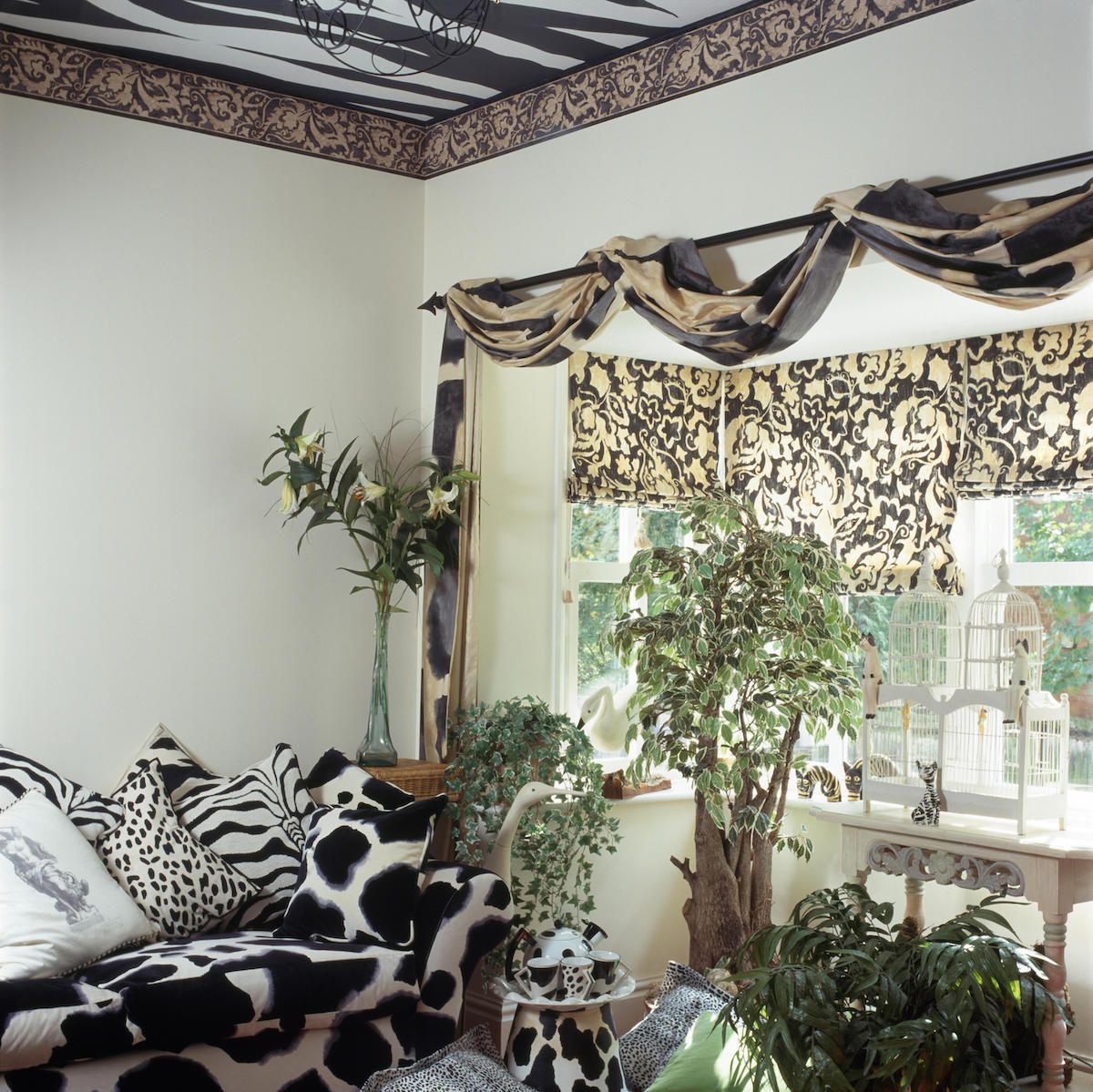 Melns + balts dzīvnieku dīvāns un spilveni deviņdesmito gadu dzīvojamā istabā ar drapētu audumu uz staba virs loga