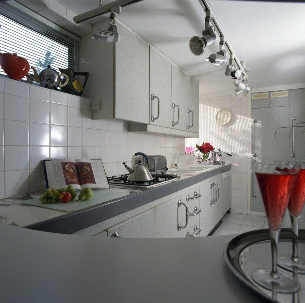 Проследете прожекторите и белите плочки в кухнята от деветдесетте с бледосиви вградени модули и чайник Alessi на плота