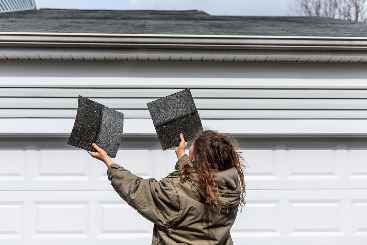 Majitelka domu stojí před domem a drží dva šindele střešních tašek, které kontrolují poškození