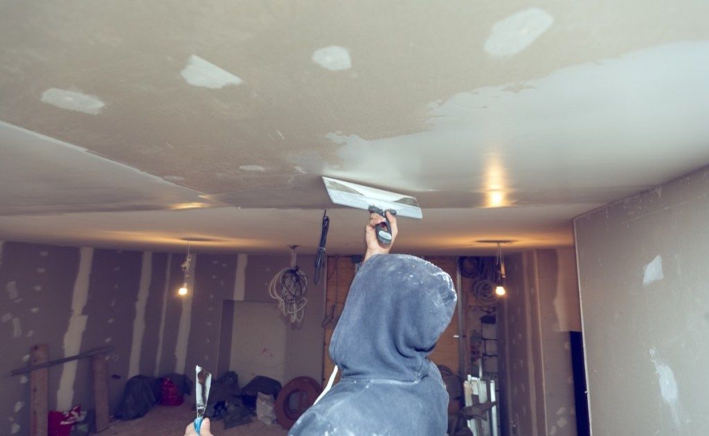 persoană în glugă cu glugă acoperind un tavan de gips-carton într-o cameră neterminată