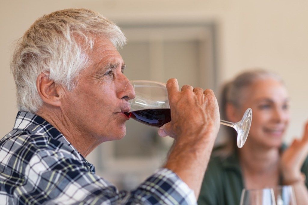 Sretni stariji čovjek koji pije čašu crnog vina za vrijeme ručka. Starac koji uživa u vinu s prijateljima u pozadini. Izbliza lice aktivnog i zdravog starijeg muškarca koji kuša vino.