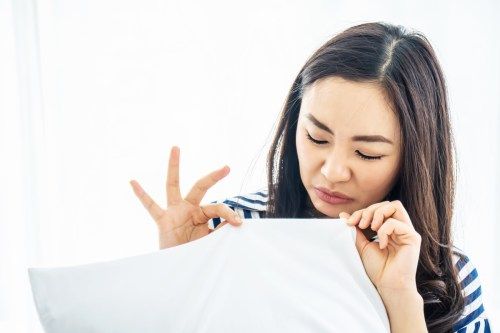 Graži moteris uostydama baltą pagalvę su bjauriu ar ūksmingu kvapu, veidas rodo bjaurius simptomus baltame miegamajame, idėja žmonėms ir švarus čiužinys