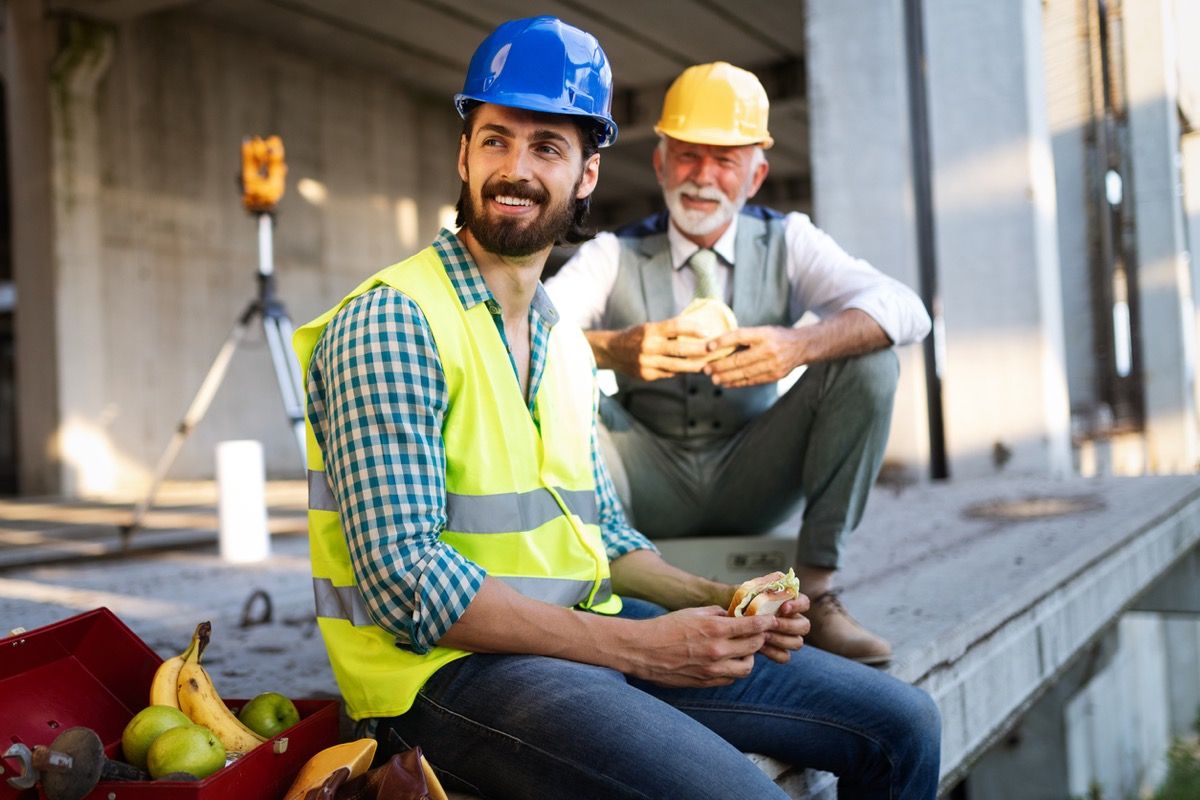 yngre og eldre bygningsarbeider som spiser smørbrød og smiler