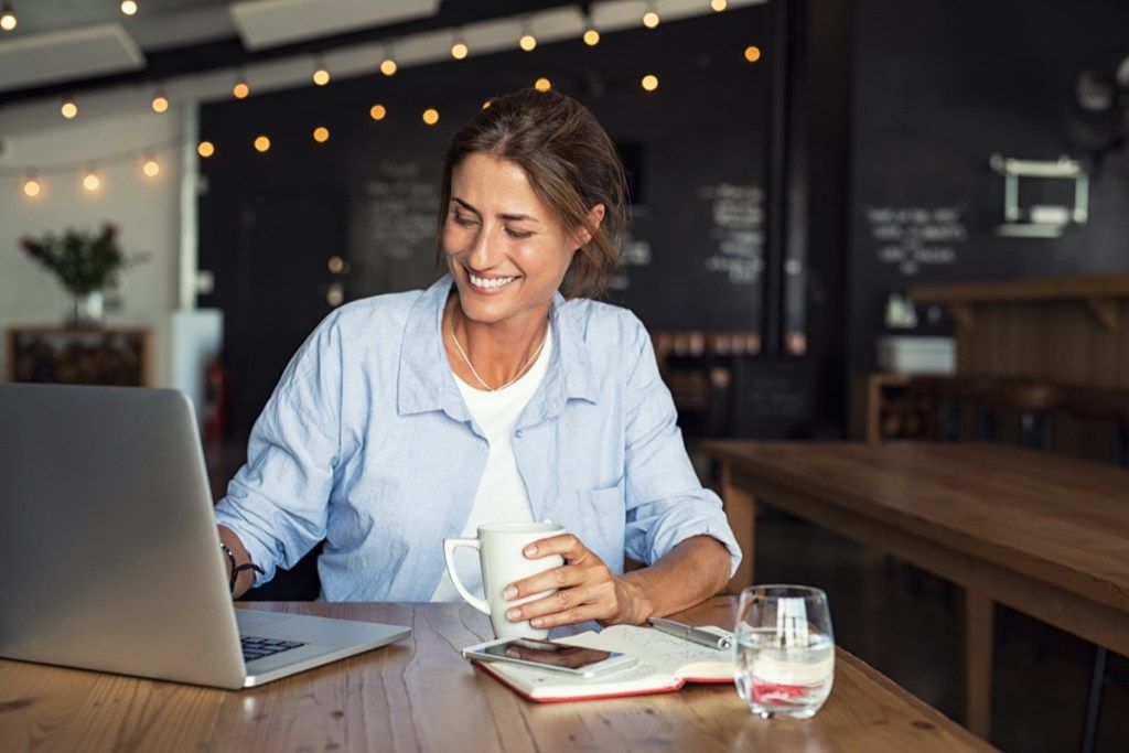 Moteris laiminga ir besišypsanti dirbdama su nešiojamuoju kompiuteriu iš kavinės