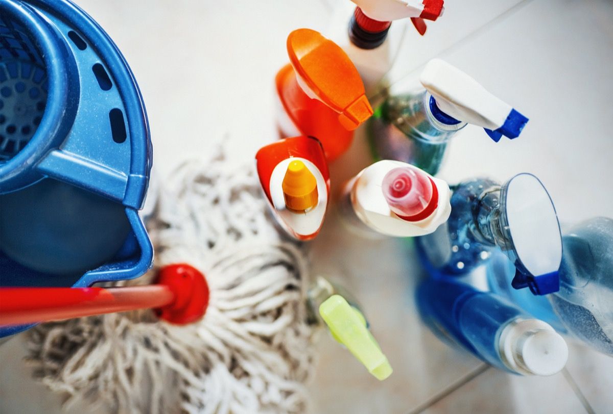 Nærbillede ovenfra af uigenkendelige rengøringsprodukter til hjemmet med blå spand og en moppe på siden. Alle produkter anbragt på hvidt flisebelagt badegulv.