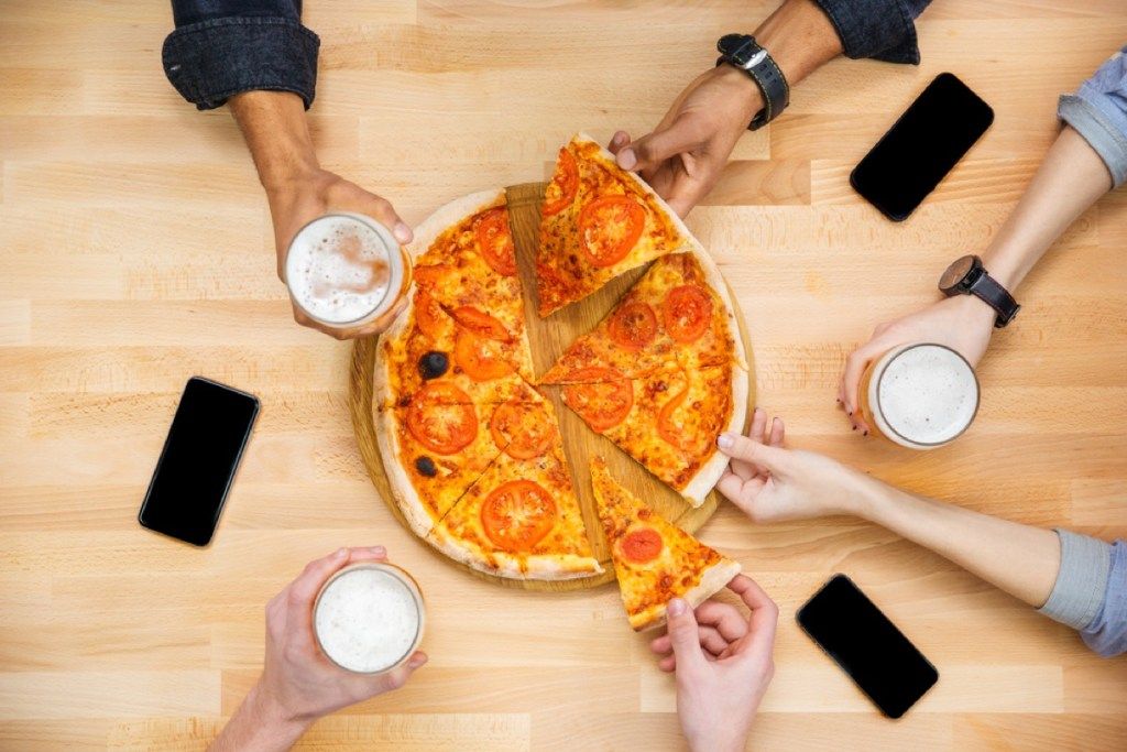 kezek pizzaszeletekért nyúlnak az asztalon, telefonokkal és korsó sörrel
