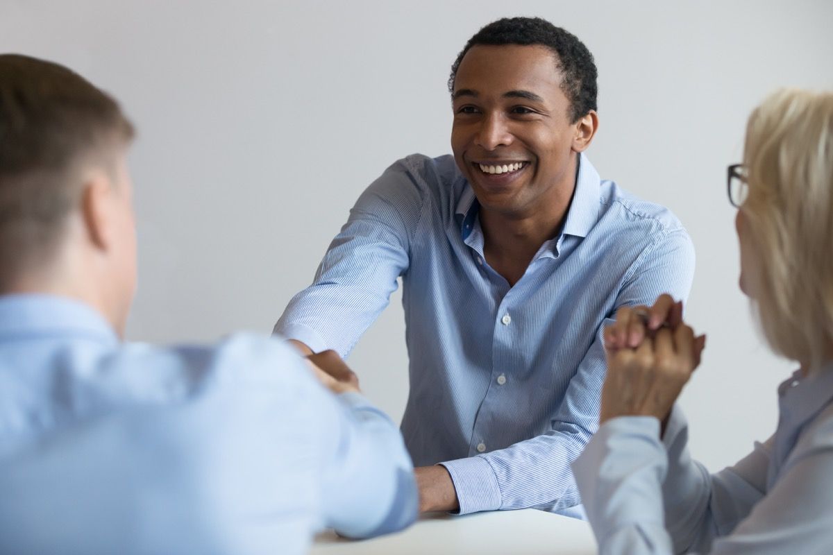 Homem negro de 40 e poucos anos aperta a mão de uma entrevista com um homem branco sentado à mesa, etiqueta acima dos 40
