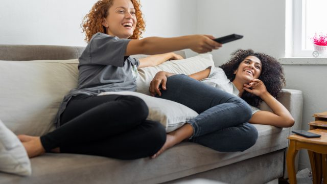   Две млади жени, седнали на диван с дистанционно и гледащи телевизия