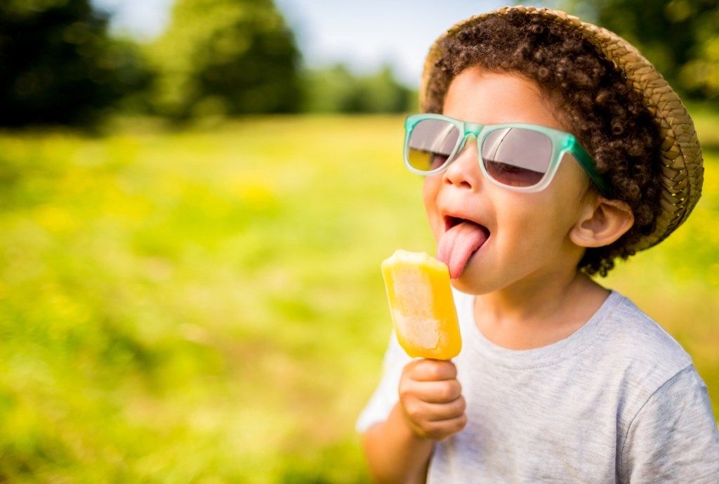 A língua de um menino com cabelo encaracolado está tocando uma guloseima congelada de laranja. Ele está vestindo uma camiseta cinza, um chapéu de palha e óculos de aro azul. O fundo é desfocado e consiste em um campo aberto de grama verde com árvores e arbustos à distância.