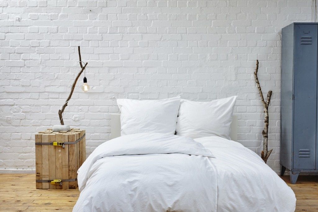 Cama blanca en el piso con pared de ladrillo blanco detrás y mesita de noche de madera