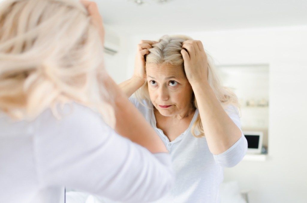 vecāka sieviete skatās uz matiem spogulī, izskatās labāk pēc 40