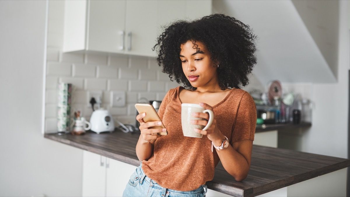 Bidikan wanita muda menggunakan ponsel cerdas dan minum kopi di dapur di rumah