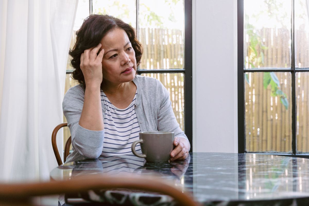 Zdenerwowana starsza kobieta przeczesuje dłonią włosy, patrząc przez okno swojego domu. Siedzi przy kuchennym stole. Przed nią stoi filiżanka kawy.