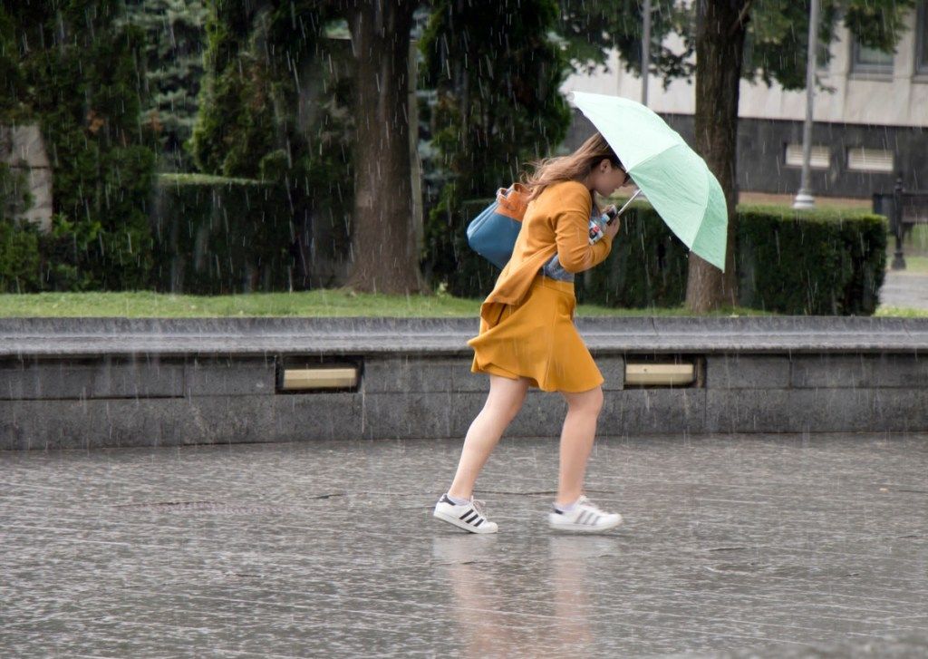 बेलग्रेड, सर्बिया - 14 जून, 2018: शहर के पार्क में अचानक भारी और तेज़ वसंत की बारिश में छतरी के नीचे एक युवती, पानी की बोतल पकड़े