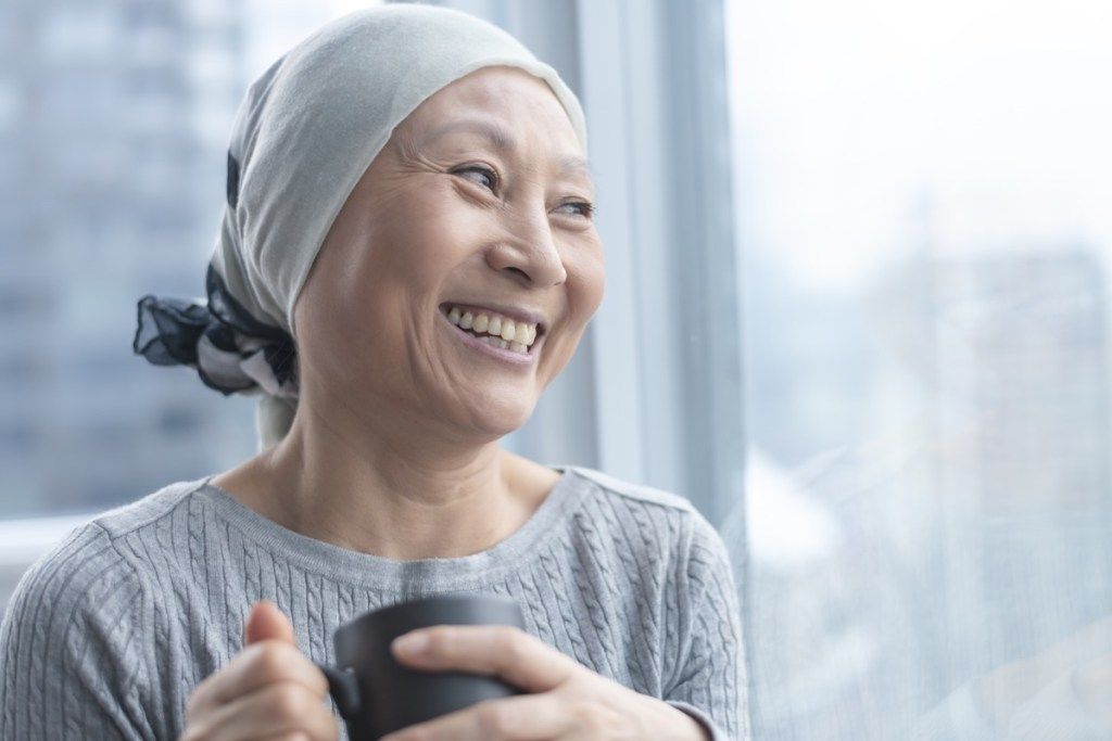 Korealainen vanhempi nainen, jolla on syöpä, on päällään huivi. Hän seisoo ja pitää kupin teetä. Nainen nojaa ikkunaa vasten ja hymyilee kiitollisuudella ja toivolla.