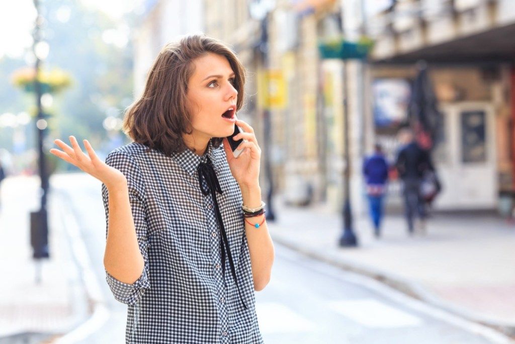 Pykusi moteris gatvėje kalba išmaniuoju telefonu. Dėvi laisvalaikio drabužius.