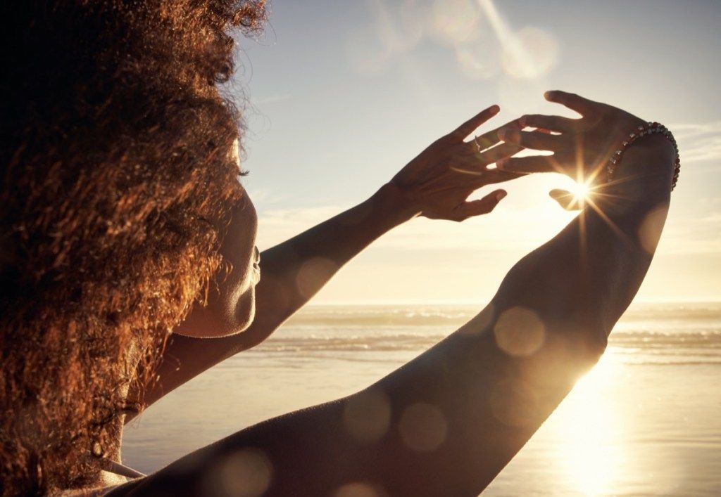 ساحل سمندر پر اپنے ہاتھوں سے سورج کی روشنی کو روکتی ہوئی ایک نوجوان عورت کا ریرویو شاٹ