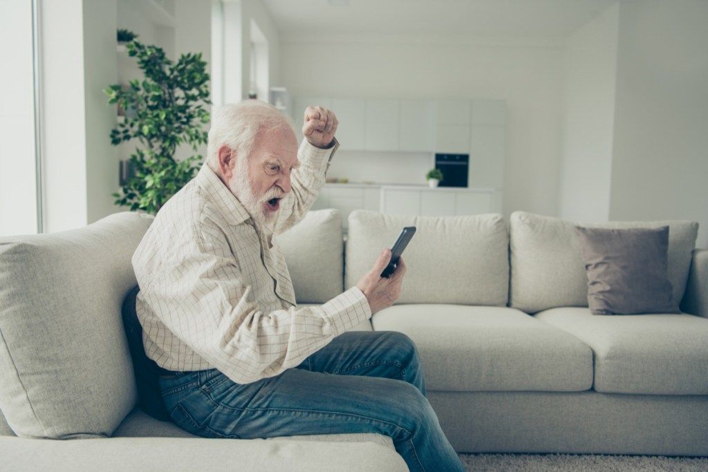 Профилен страничен изглед портрет на луд стилен старец, носещ чекирана риза, седнал на диван, държащ в ръка телефон, полудял, възмутен в бяла светлина, модерен интериорен студио