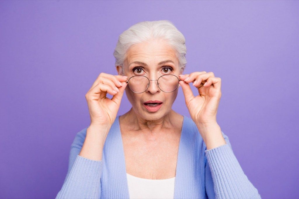 Портрет на красива приятно изумена сива коса на старица, носеща ежедневни и очила, показващ жест, слагащ очила. Изолирани над пастелно виолетово лилав фон