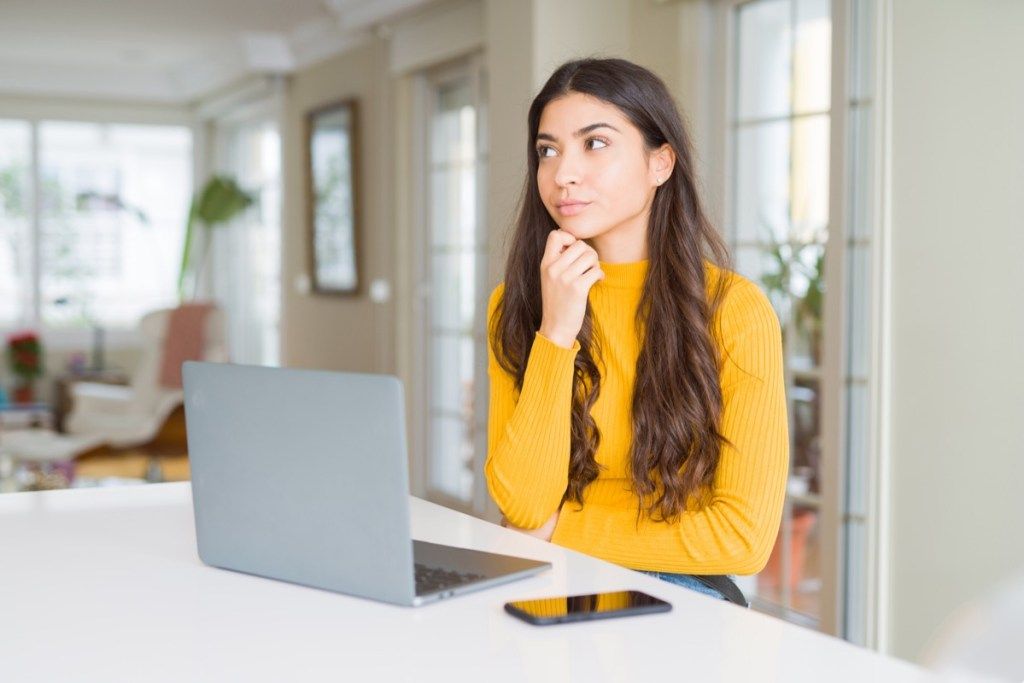 Mujer joven con ordenador portátil con la mano en la barbilla pensando en pregunta, expresión pensativa. Sonriendo con rostro pensativo. Concepto de duda.