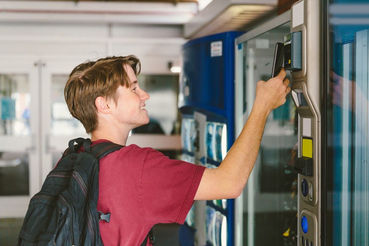 Dospívající chlapec studenta s batohem používá mobilní telefon k platbě za občerstvení a pití v automatu