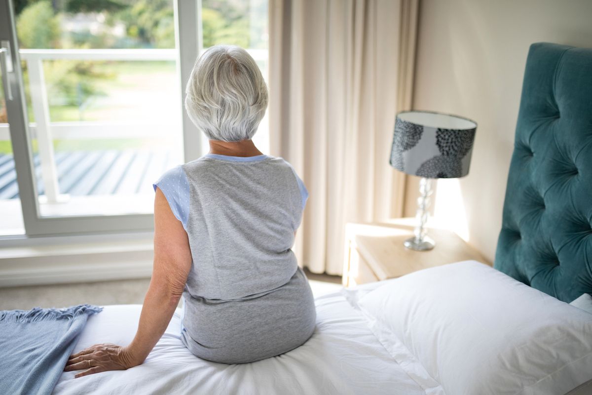 Zadní pohled na starší ženu sedící na posteli v ložnici, při pohledu z okna