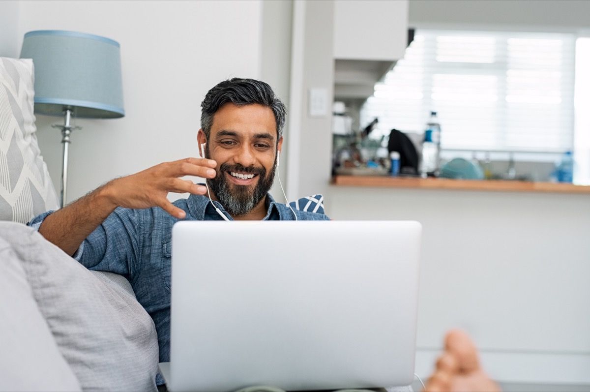איש של גיל העמידה של צבע נרגע על הספה בזמן שיחות וידאו באמצעות מחשב נייד בבית