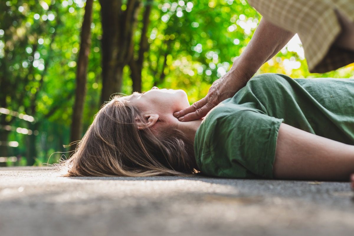 medvetslös svimmad tjej som får en puls kontrollerad av en gammal kvinna - Tonåring som ligger på marken medan hennes puls verifieras av en äldre medborgare på tonåring