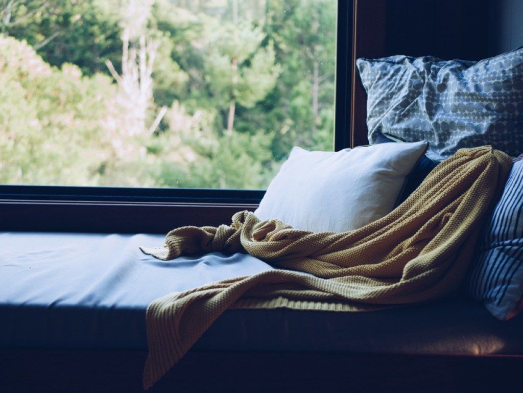 кът за четене с две сини възглавници, жълто хвърляне и бяла възглавница пред прозореца