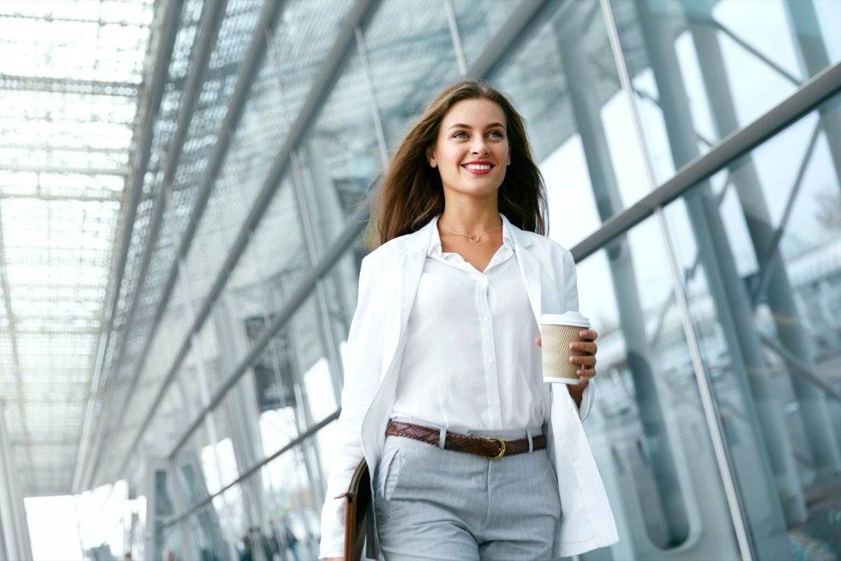 Жена отива на работа с кафе, разхождайки се в близост до офис сграда. Портрет на успешна бизнес жена, която държи чаша гореща напитка в ръка по пътя си към работа на улицата City Street