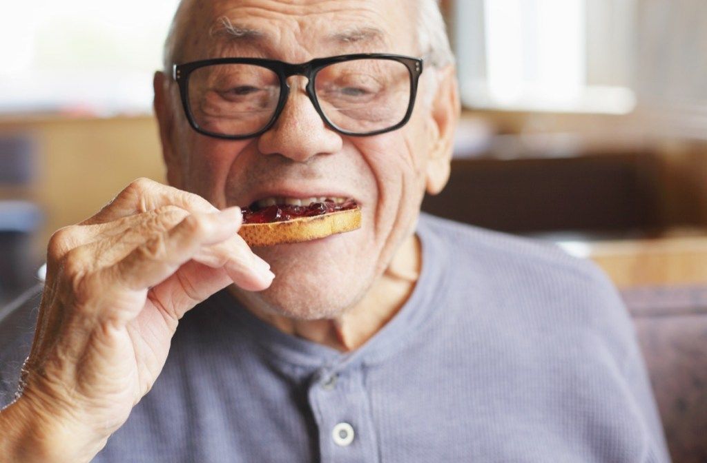 Възрастен мъж се наслаждава на хапка от препечения си хляб, намазан със сладко желе, конфитюр по време на закуска в ресторант.