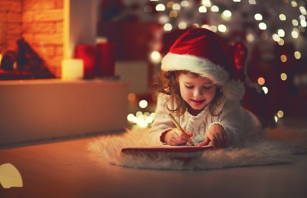 mažas vaikas priešais eglutę rašo Kalėdų Seneliui laišką
