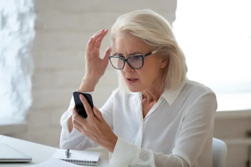  Una anciana con gafas y sentada en un escritorio mira su smartphone con una mirada sorprendida y confundida, tal vez víctima de una estafa.