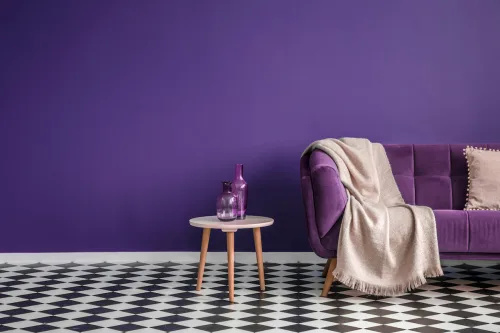   Mörklila soffa med en filt bredvid ett litet bord med flaskor som står på svartvitt rutgolv i ett minimalistiskt vardagsrum med lila väggar.