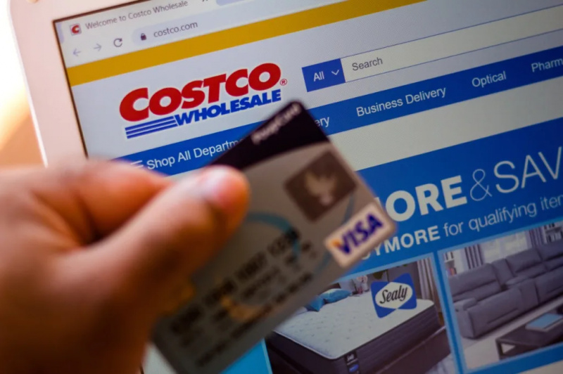   कॉस्टको होलसेल कॉर्पोरेशन की वेबसाइट पृष्ठभूमि में एक लैपटॉप पर दिखाई गई है और एक व्यक्ति के पास बैंक कार्ड है