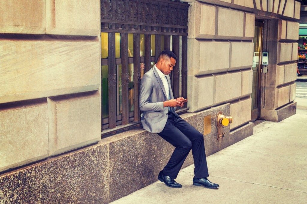 アフリカ系アメリカ人の男性が読書、路上で携帯電話でテキストメッセージを送信、旅行、ニューヨークでの作業、灰色のブレザー、黒のズボン、革の靴を履いて、ヴィンテージの窓枠に座っています。 Instagramのフィルタリングされた外観-画像