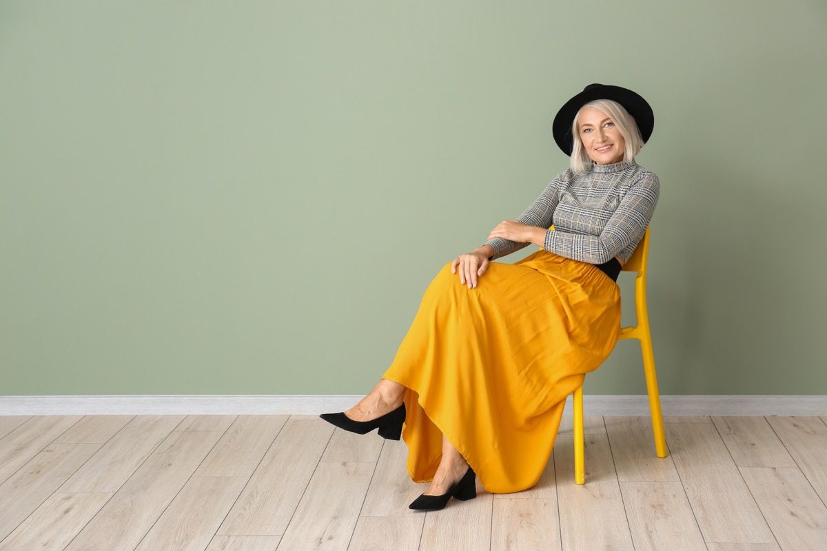 40-ies metų moteris, sėdinti ant kėdės, vilkinti drąsiai ryškiai geltonos spalvos sijono aprangą su kepure