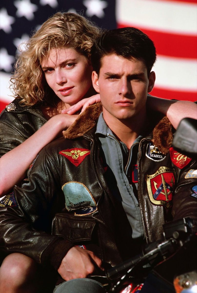 Tom Cruise con chaqueta de bombardero con Kelly McGillis recostada sobre su espalda en la película Top Gun, Cruise lleva una