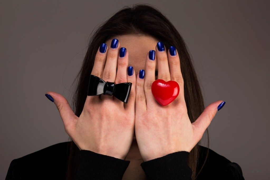 काले धनुष की अंगूठी और लाल दिल की अंगूठी पहने एक महिला अपने हाथों से अपना चेहरा ढँक लेती है