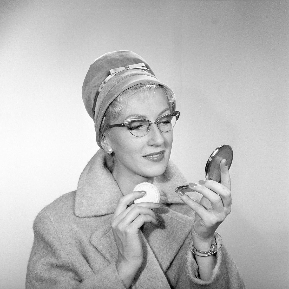 Machiajul anilor 1950. O tânără se privește în oglinda de buzunar și își îmbunătățește machiajul. Poartă o pălărie la modă, ochelari tipici anilor 50 și o haină. Anii 1950