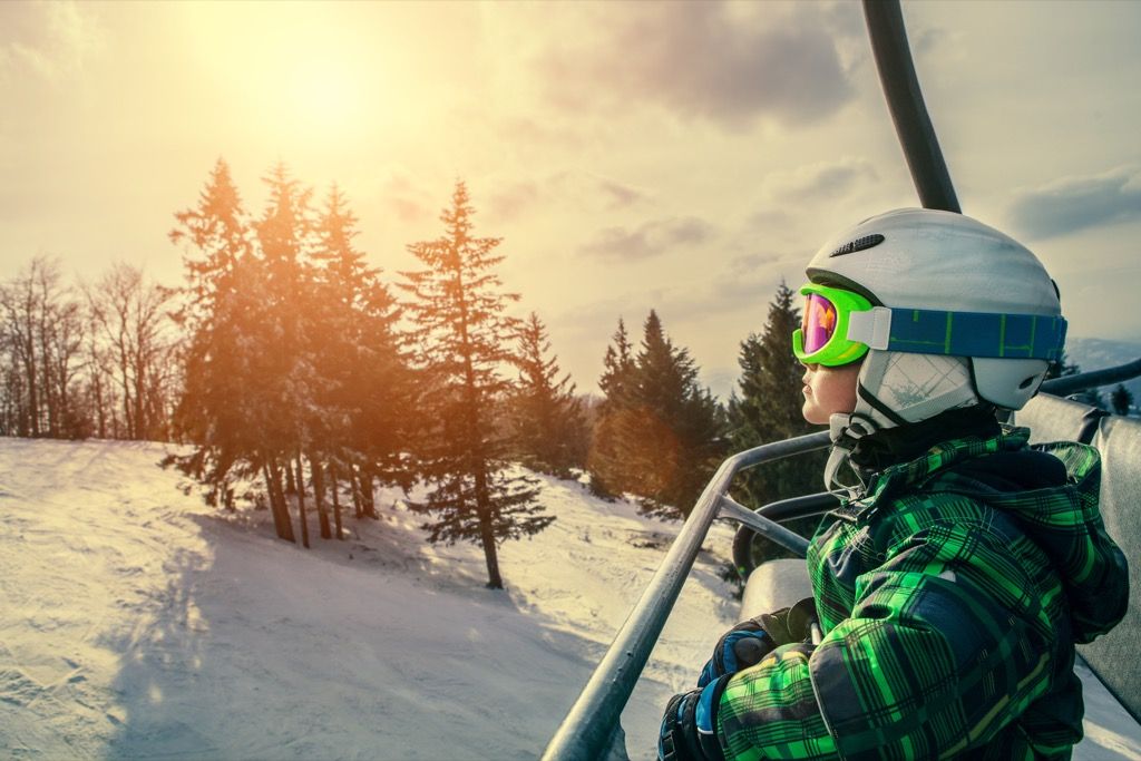 헬멧, 고글, 스키트 코트를 입은 어린 소년이 눈 덮인 산을 뒤로하고 의자 리프트에 앉아 있습니다.